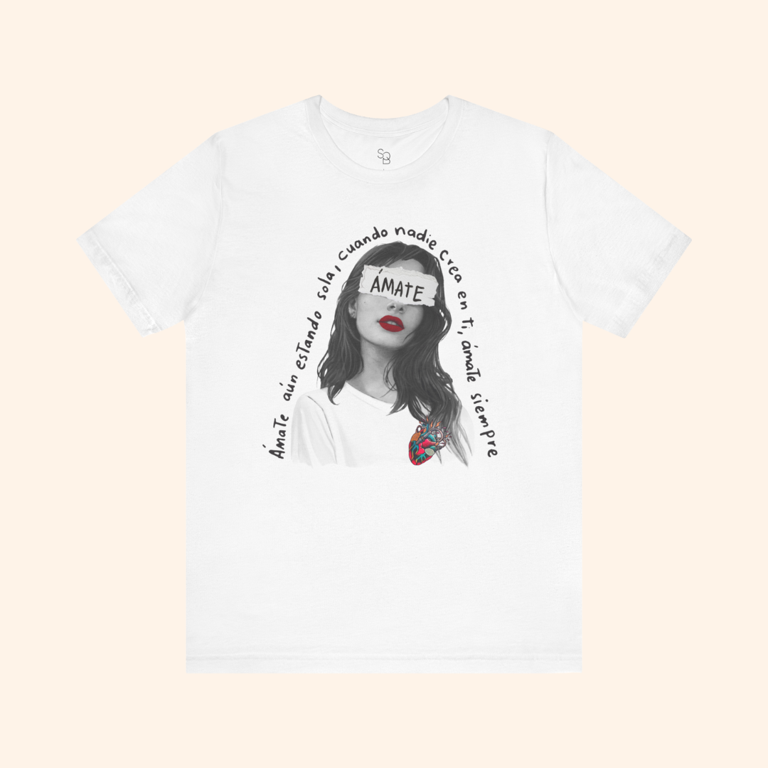 Flat lay of white Amate t-shirt on neutral background showing artistic female portrait, red lips, anatomical heart illustration, and Spanish self love quote.