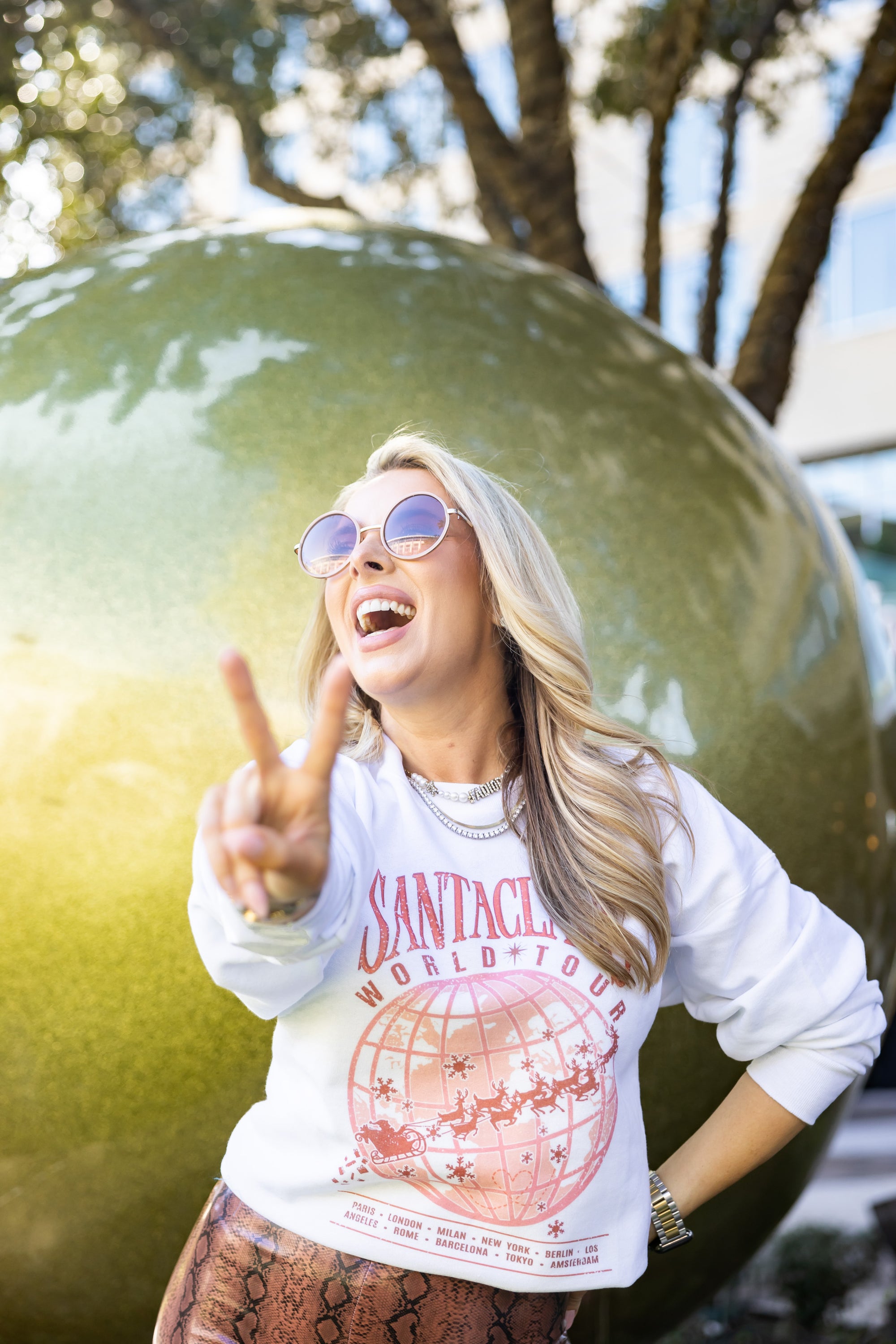 Model outdoors wearing Santas World Tour sweatshirt, lifting sunglasses, festive backdrop with oversized holiday ornament, bold Christmas street style look for women.
