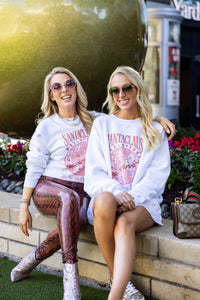 Two women sitting outdoors wearing white Santas World Tour sweatshirts with red tour map design, styled with sunglasses, sparkly boots, and designer bag, casual festive street style look for holiday season.