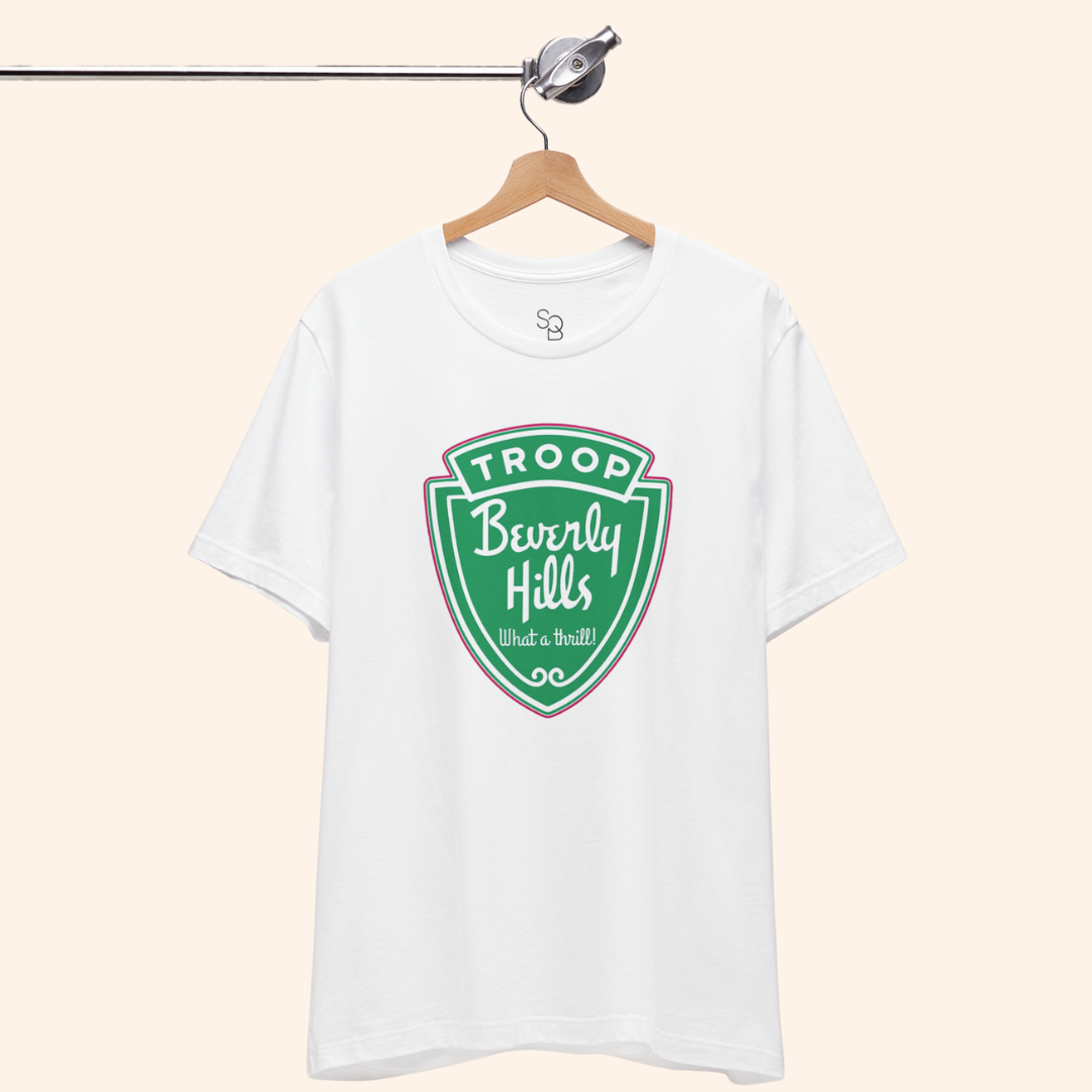 Troops T-Shirt hanging neatly from a wooden hanger, white cotton fabric with a bold green badge graphic centered on the chest.