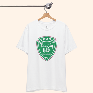 Troops T-Shirt hanging neatly from a wooden hanger, white cotton fabric with a bold green badge graphic centered on the chest.