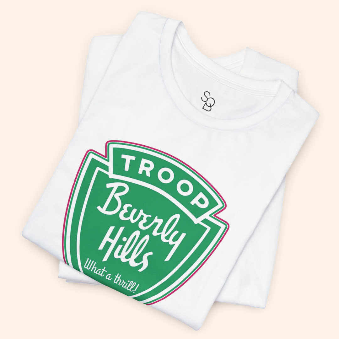 Folded white Troops T-Shirt with the green retro badge graphic visible on top, crisp cotton texture, boutique style presentation.
