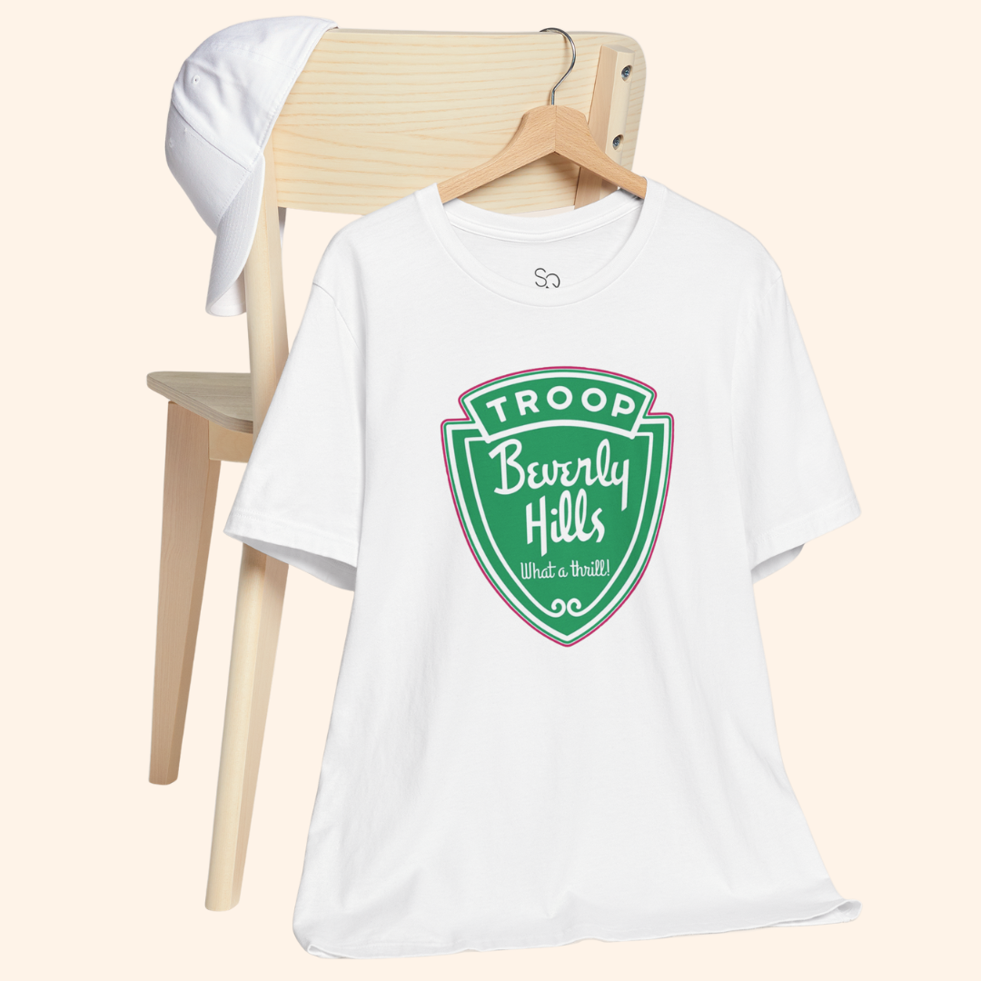 White Troops graphic tee hanging on a wooden chair next to a white cap, showcasing the green retro badge design and relaxed fit.