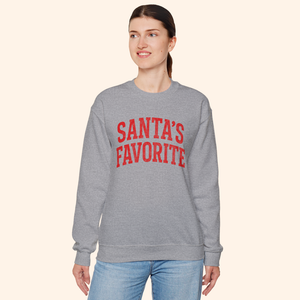 Full view of a woman wearing the Santas Favorite graphic sweatshirt with a comfortable unisex fit. Heather gray fleece with red lettering styled for an everyday holiday outfit that blends comfort and seasonal fashion.