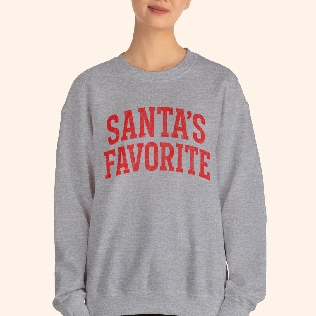A woman wearing a heather gray Santas Favorite sweatshirt with bold red lettering, standing against a neutral backdrop. Relaxed unisex fit with casual styling that highlights the holiday graphic pullover and its soft winter fleece texture.