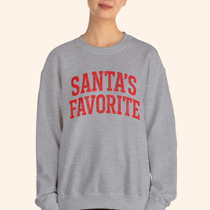 A woman wearing a heather gray Santas Favorite sweatshirt with bold red lettering, standing against a neutral backdrop. Relaxed unisex fit with casual styling that highlights the holiday graphic pullover and its soft winter fleece texture.