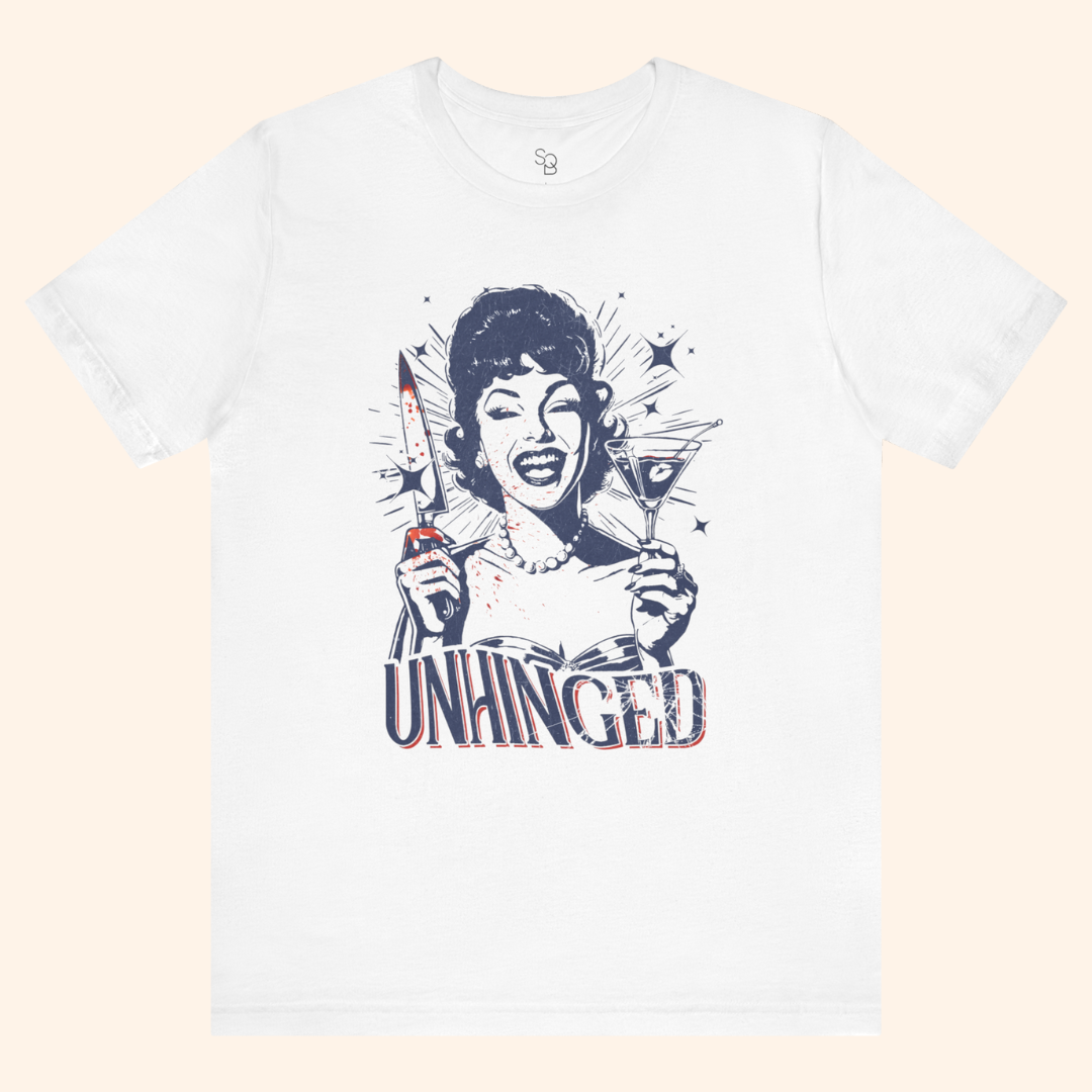 Full front flat lay of the Unhinged T-Shirt on a soft beige backdrop, highlighting the navy and red dark humor graphic of a smiling vintage woman with knife and cocktail, unisex white graphic tee ready to style with jeans or skirts.