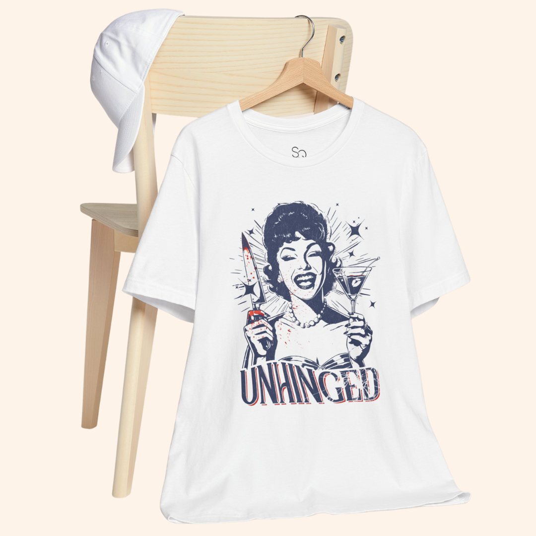 Unhinged T-Shirt hanging from a wooden chair with a white cap in the background, casual lifestyle setup that highlights the retro housewife knife and martini artwork, ideal graphic tee for off duty outfits and brunch plans.