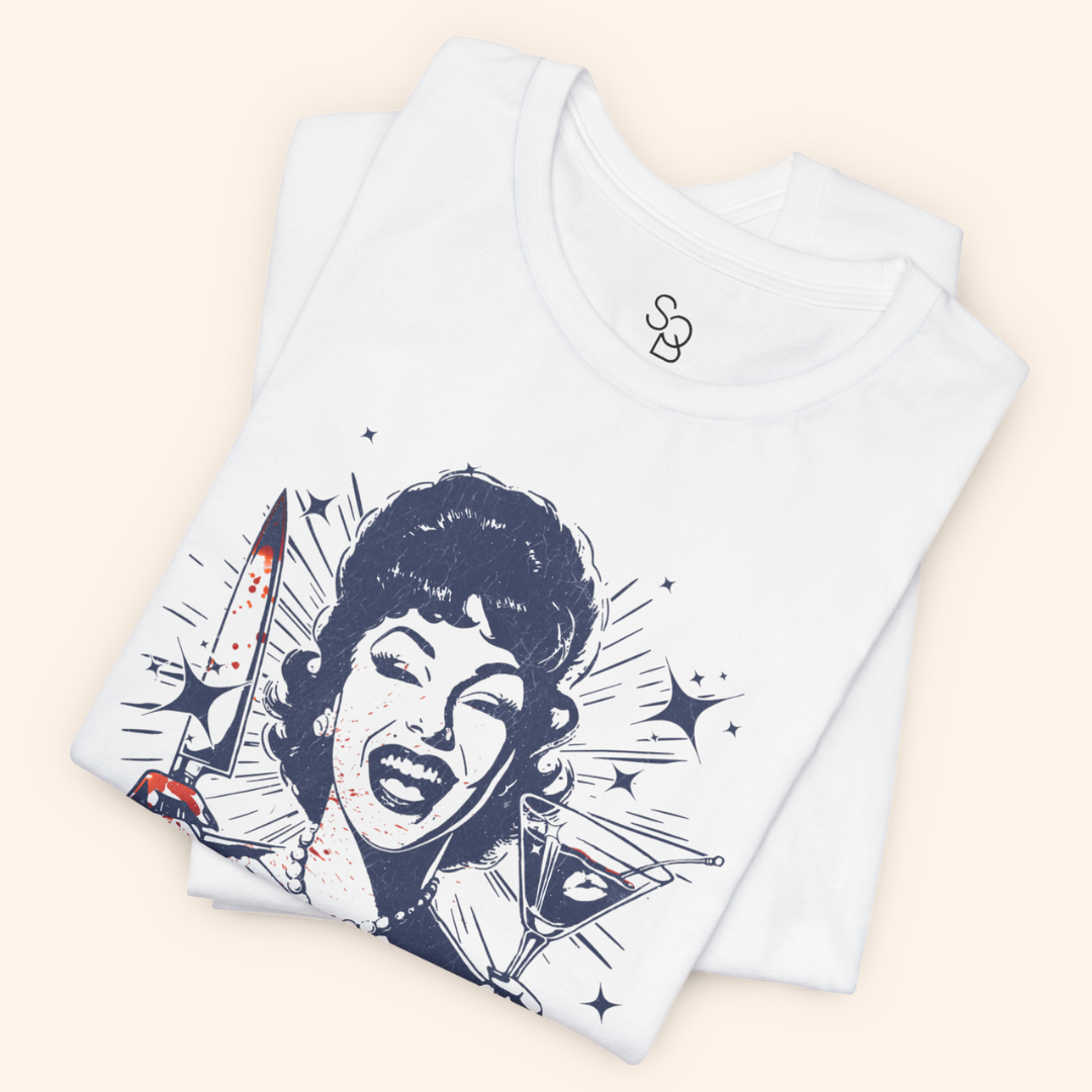 Folded white Unhinged T-Shirt stacked neatly, zoomed in on the laughing retro housewife artwork, knife and martini details visible, showcasing soft cotton fabric and bold dark humor print for gifting or everyday wear.