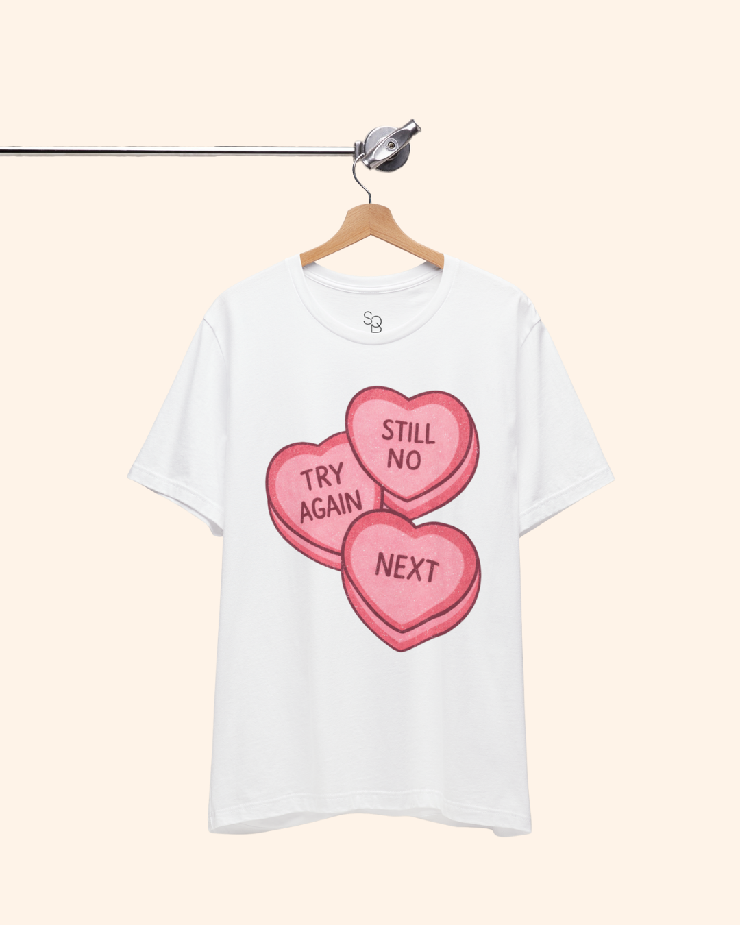 White graphic t-shirt hanging on a wooden hanger from a metal clothing rod, featuring three pink candy heart graphics that read Try Again, Still No, and Next, shown against a light neutral background.