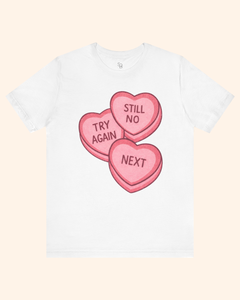 Front view of a white graphic t-shirt with three pink candy heart designs reading Try Again, Still No, and Next, centered on the chest and shown against a light neutral background.