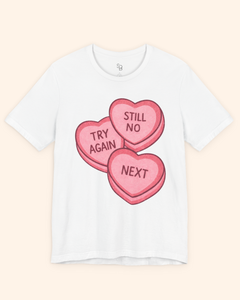 White short sleeve graphic t-shirt featuring pink candy heart artwork with the words Try Again, Still No, and Next, displayed front facing on a light beige background.