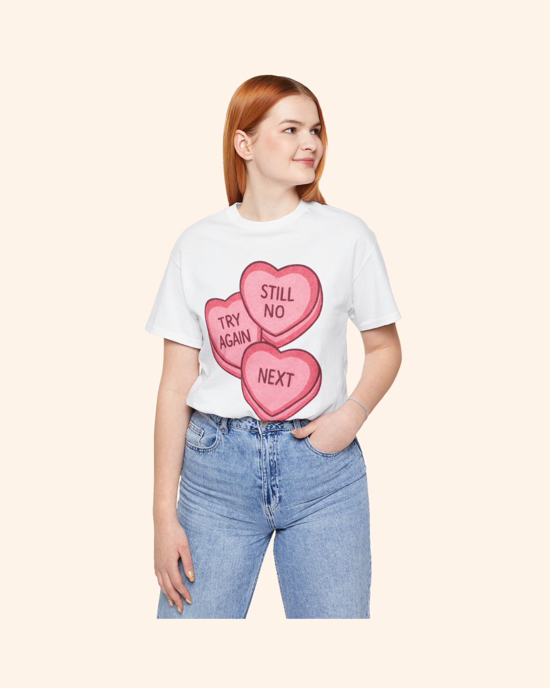 Mid-size woman wearing a white graphic t-shirt with pink candy heart artwork reading Try Again, Still No, and Next, styled casually with blue jeans and hands in pockets against a neutral background.