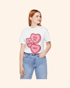 Mid-size woman wearing a white graphic t-shirt with pink candy heart artwork reading Try Again, Still No, and Next, styled casually with blue jeans and hands in pockets against a neutral background.