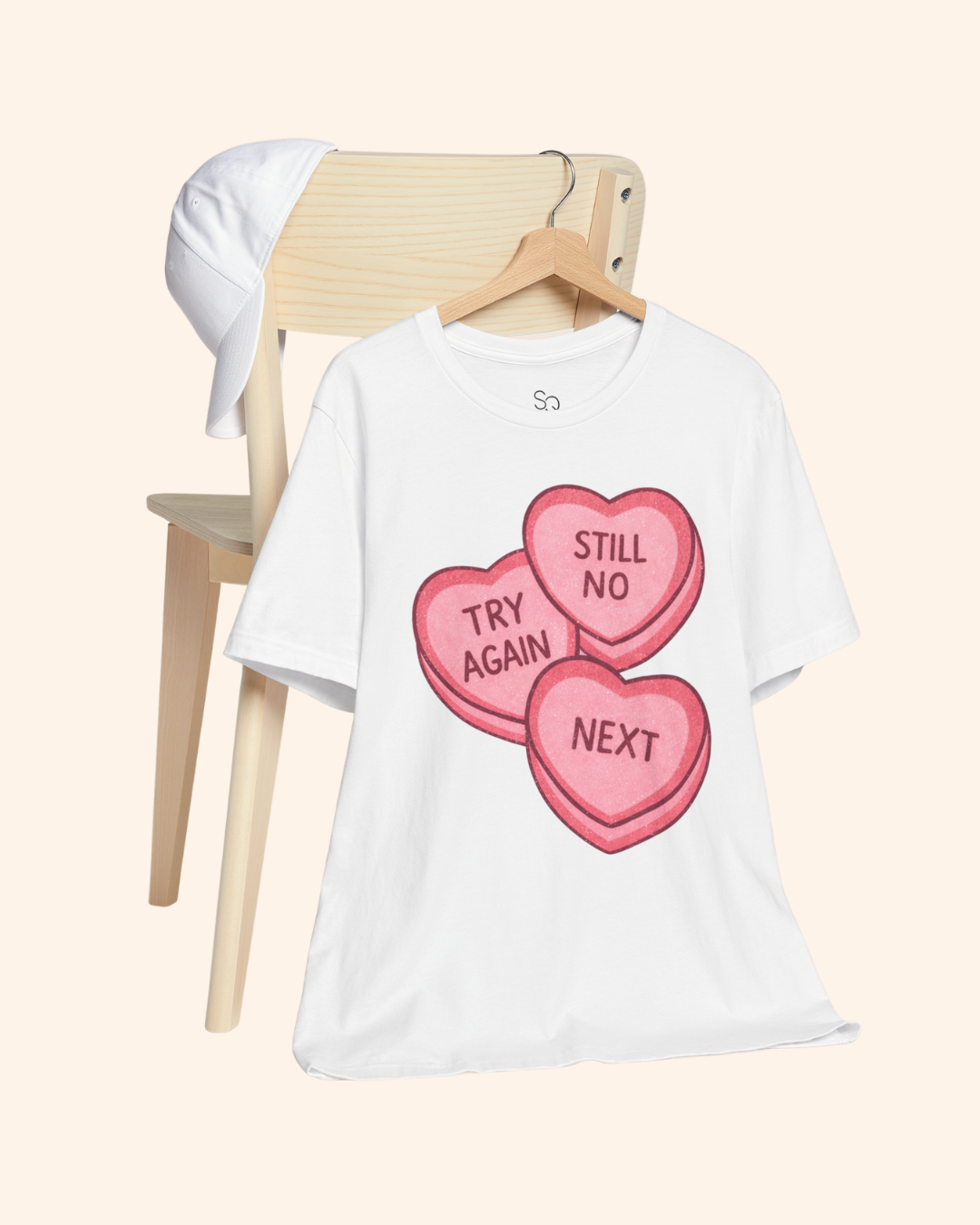 White graphic t-shirt with pink candy heart design reading Try Again, Still No, and Next, hanging on a wooden chair with a white hat placed on the chair back, styled against a soft neutral background.
