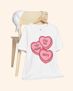 White graphic t-shirt with pink candy heart design reading Try Again, Still No, and Next, hanging on a wooden chair with a white hat placed on the chair back, styled against a soft neutral background.