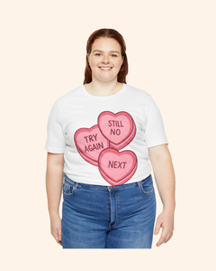 Plus size woman wearing a white graphic t-shirt with three pink candy heart designs reading Try Again, Still No, and Next, paired with blue jeans and photographed front facing against a light neutral background.