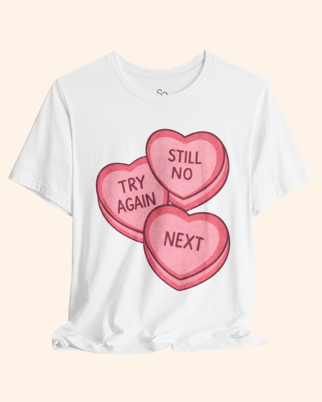White graphic t-shirt displayed flat with a soft draped hem, featuring three pink candy heart graphics that read Try Again, Still No, and Next, shown on a neutral beige background.