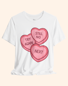 White graphic t-shirt displayed flat with a soft draped hem, featuring three pink candy heart graphics that read Try Again, Still No, and Next, shown on a neutral beige background.