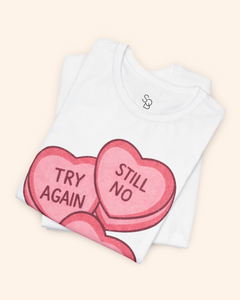 Folded white graphic t-shirt showing close detail of the pink candy heart design and neckline, with the words Try Again and Still No visible on the top fold.