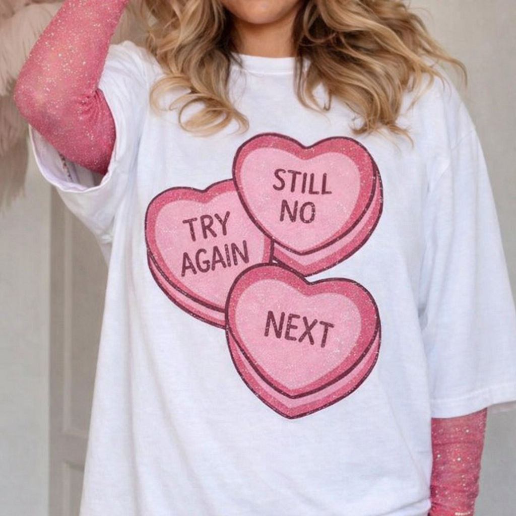Woman wearing a white graphic t-shirt with three pink candy heart designs reading Try Again, Still No, and Next. Shirt is styled layered over a pink long sleeve top, shown on a real model from chest to waist against an indoor background.