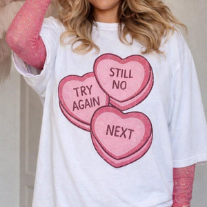 Woman wearing a white graphic t-shirt with three pink candy heart designs reading Try Again, Still No, and Next. Shirt is styled layered over a pink long sleeve top, shown on a real model from chest to waist against an indoor background.