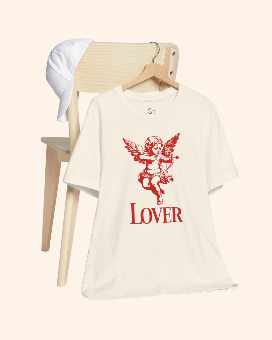 Natural color graphic t shirt hanging on a wooden chair with red cupid cherub design and lover text centered on the chest.