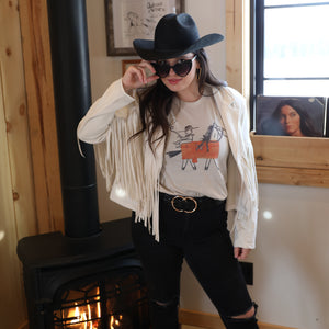 Western graphic tee paired with fringe jacket, cowgirl hat, and black denim for bold rodeo fashion.

