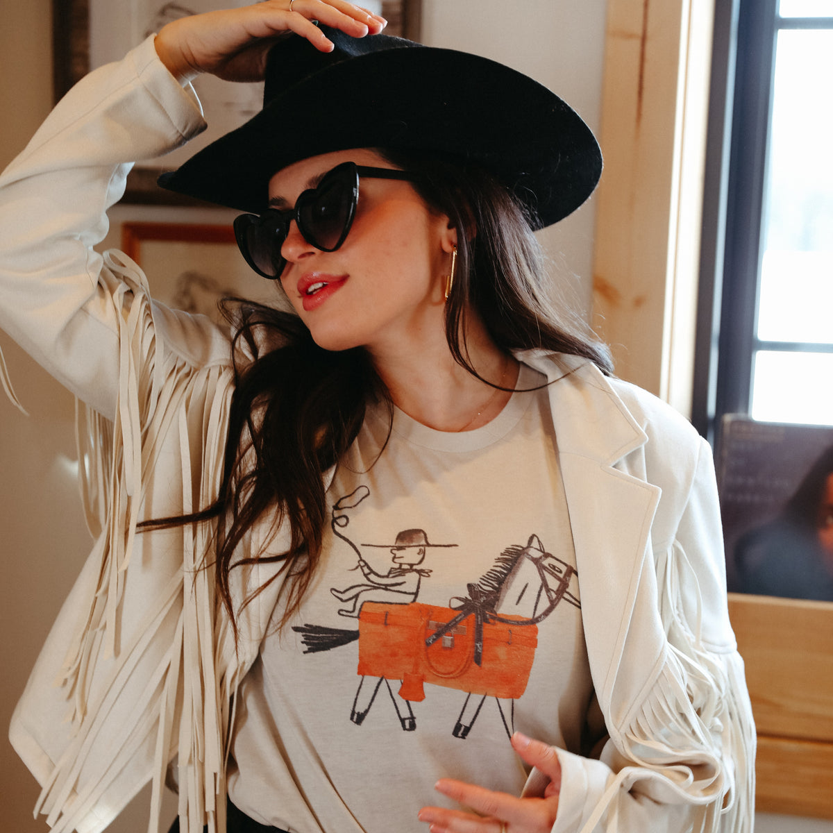 Western graphic tee paired with fringe jacket, cowgirl hat, and black denim for bold rodeo fashion.

