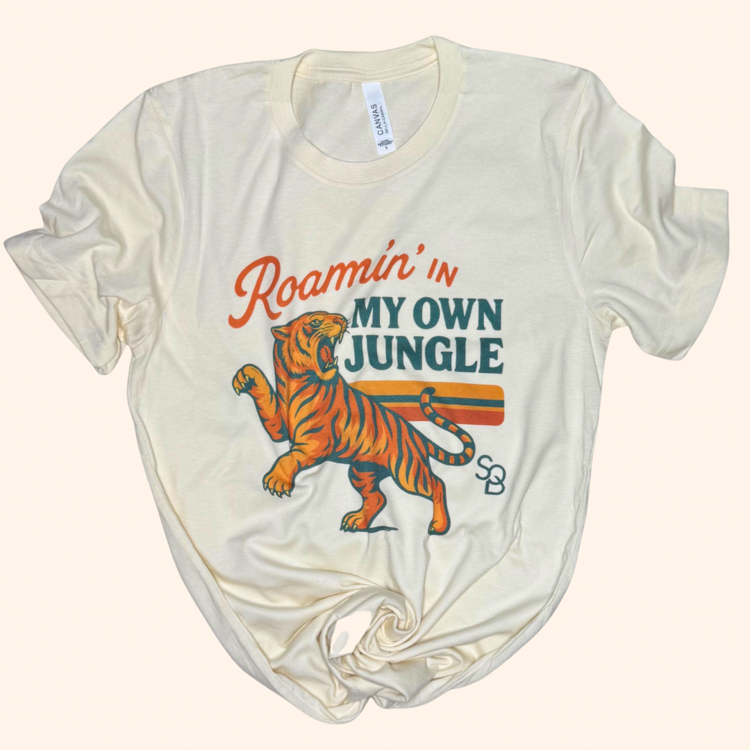 Beige tiger travel tee laid flat with visible graphic reading “Roamin’ in my own jungle” on front center.

