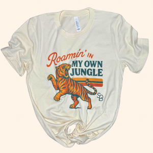 Beige tiger travel tee laid flat with visible graphic reading “Roamin’ in my own jungle” on front center.

