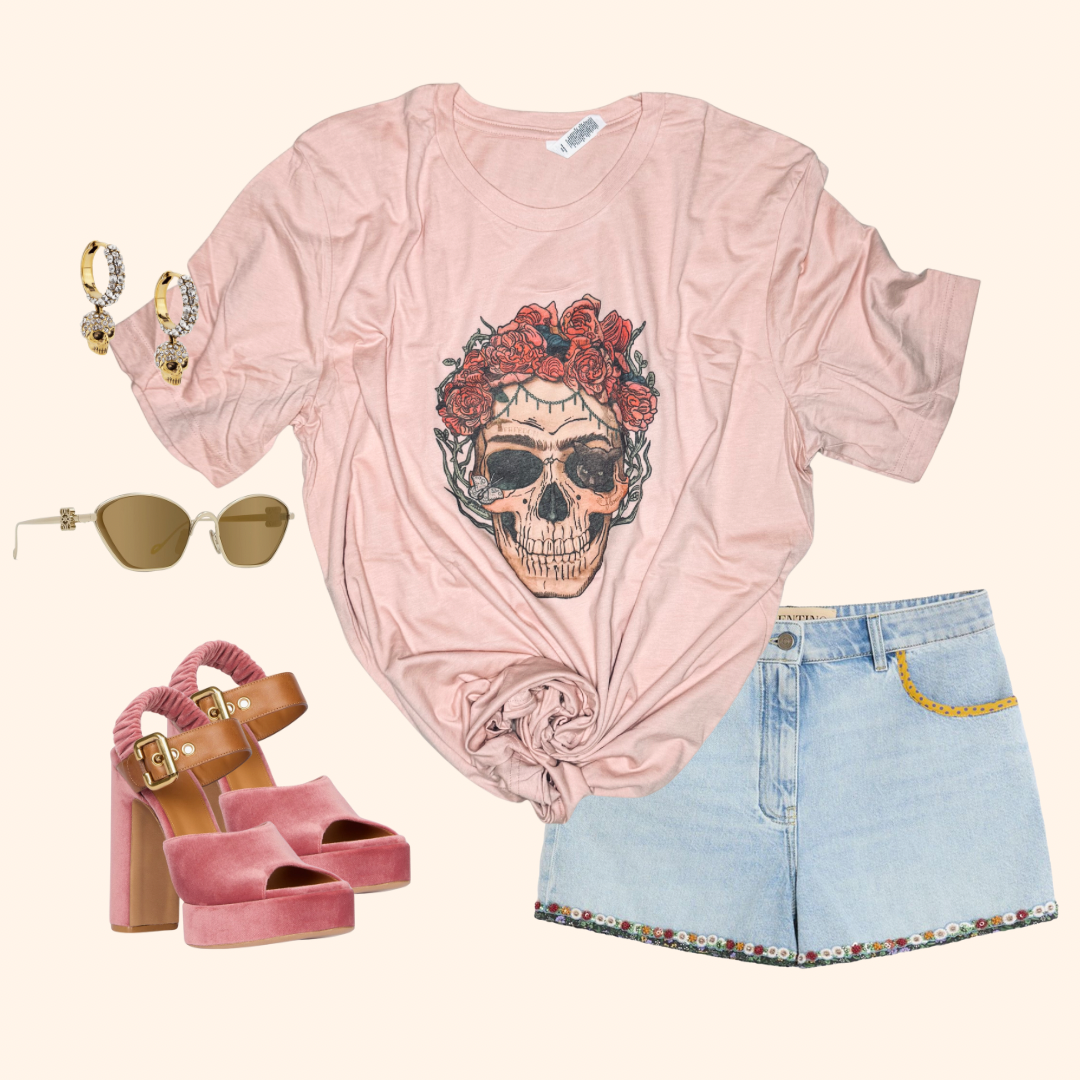 Flat lay of peach graphic tee featuring a skull with red roses, paired with light wash embroidered denim shorts, chunky pink velvet heels, gold skull hoop earrings, and cat-eye sunglasses with gold detailing.

