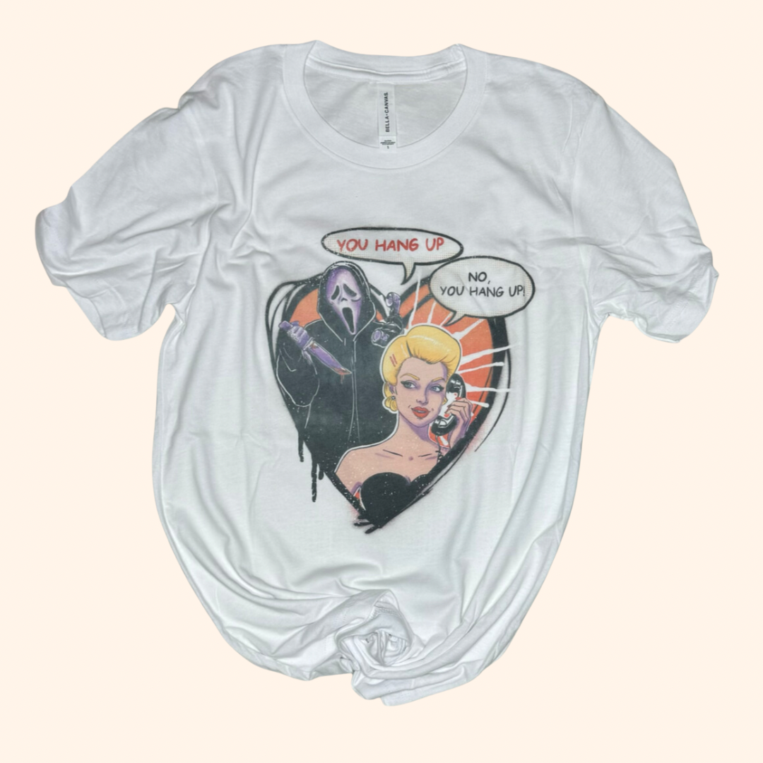 Flat lay of Hang Up Tee featuring retro cartoon-style horror parody artwork. Shows a blonde woman on the phone with a masked figure behind her in a comic book panel. Printed on a white unisex Bella Canvas tee. Laid flat to show true shape, soft texture, and design clarity.
