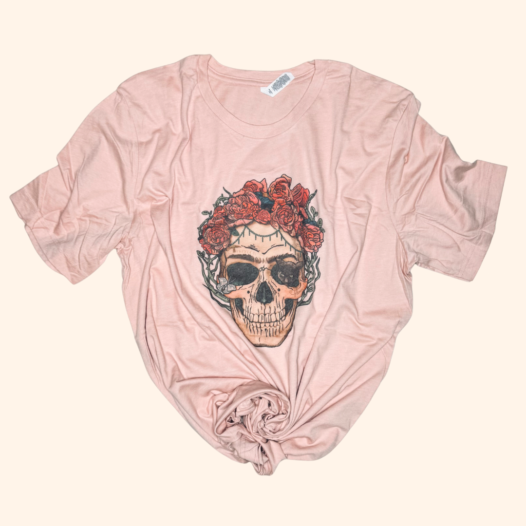Peach graphic tee featuring a detailed skull with red rose crown and vine accents, styled with a center front knot, displayed flat on a neutral beige background.

