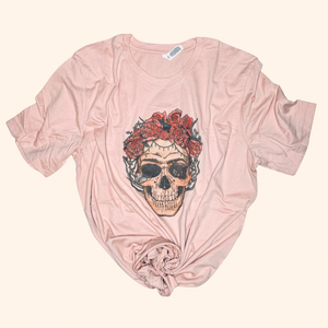 Peach graphic tee featuring a detailed skull with red rose crown and vine accents, styled with a center front knot, displayed flat on a neutral beige background.


