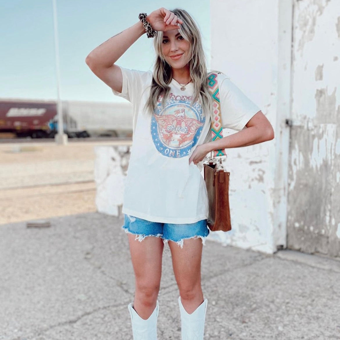 Woman wearing graphic tee with distressed denim shorts and white cowgirl boots, accessorized with a brown leather bag and western-print strap.