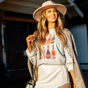 Model wears champagne bottle sweatshirt with white mini skirt, glitter jacket, and pink hat. Fun holiday or girls trip outfit.