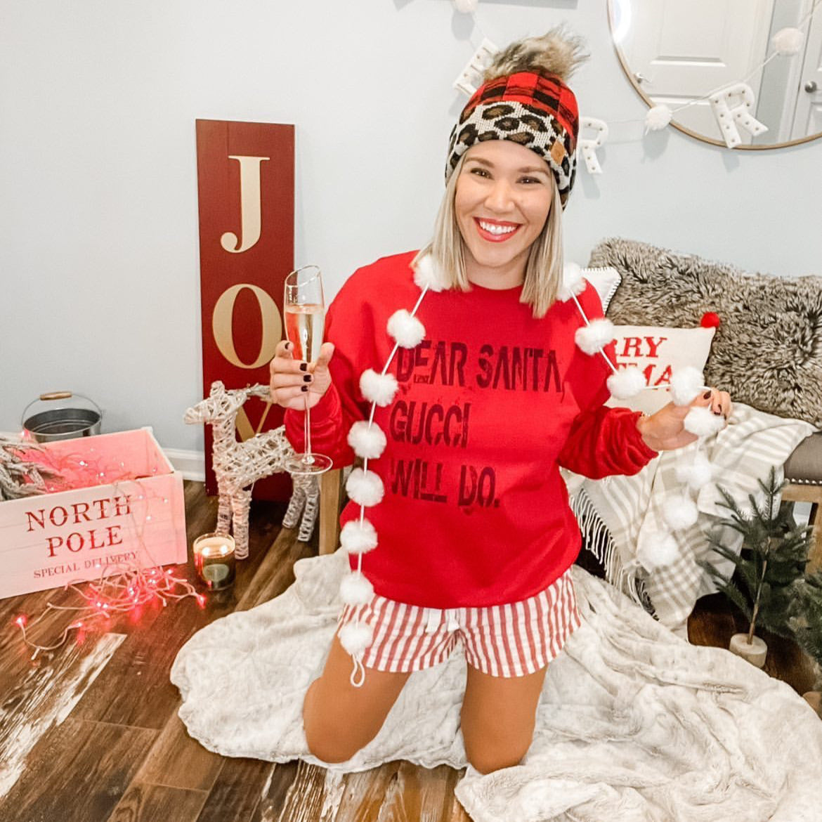 Woman wearing red Christmas sweatshirt with striped pajama shorts, tall socks, and holding a champagne glass in a cozy festive setting.

