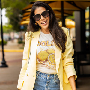 Smiling woman walking in ripped jeans and yellow blazer over retro lemon graphic tee that says “DOLCE – everyday is a fresh start” — casual street style with sunglasses and gold jewelry