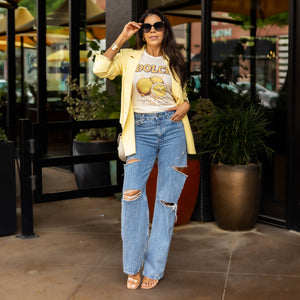 Full-body shot of woman wearing DOLCE lemon graphic tee tucked into ripped wide-leg jeans with a yellow blazer and nude heels — standing outside a café for a casual-chic summer outfit