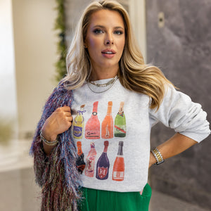 Gray sweatshirt with champagne print, styled with green pants and scarf. Playful party outfit captured inside elevator setting.

