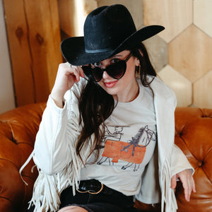 Person wearing a black cowboy hat, sunglasses, and a white fringe jacket sitting on a brown leather couch.