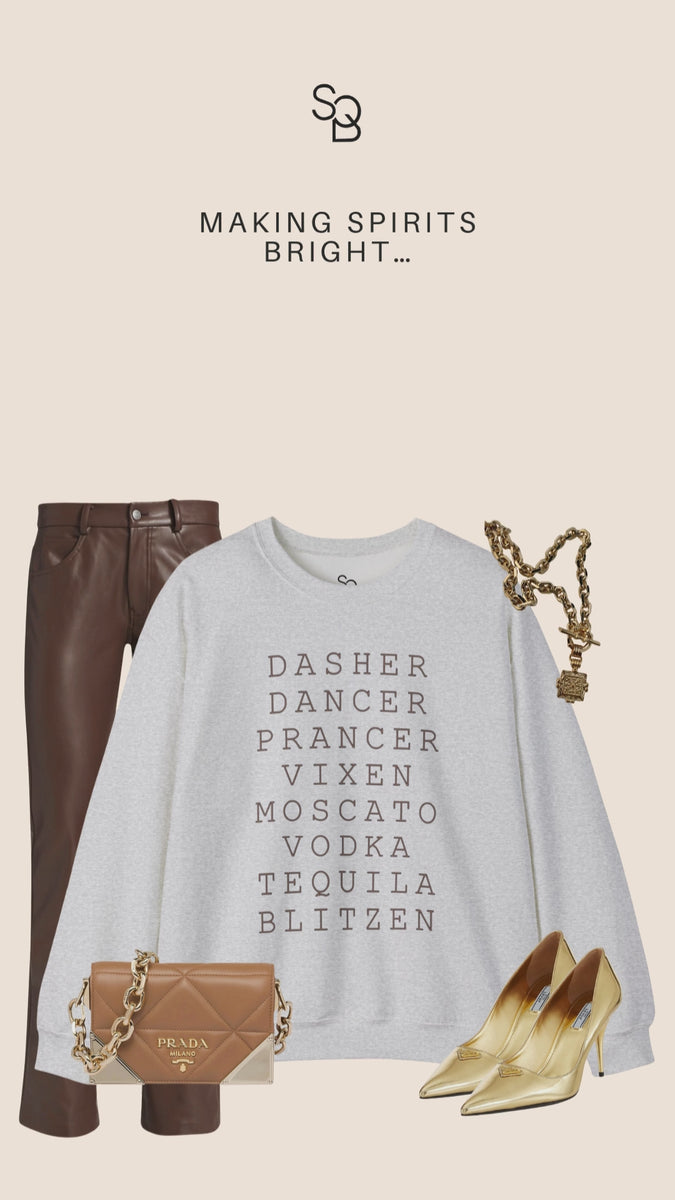 Gray Reindeer Fun sweatshirt with bold black text that lists Santa’s reindeer alongside cocktail names, styled with brown faux leather pants, gold heels, and chunky gold jewelry on a beige background, chic and witty holiday party outfit inspiration.
