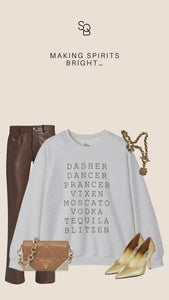 Gray Reindeer Fun sweatshirt with bold black text that lists Santa’s reindeer alongside cocktail names, styled with brown faux leather pants, gold heels, and chunky gold jewelry on a beige background, chic and witty holiday party outfit inspiration.