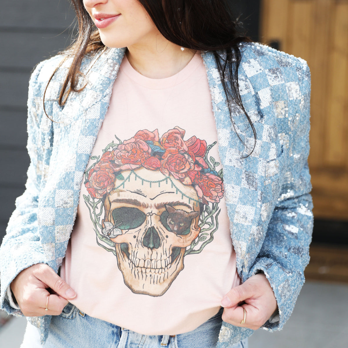 Woman wearing a peach skull graphic tee with red floral crown, styled with a sequin blue checkered blazer, pearl-studded denim headband, and high-rise light wash jeans, standing in natural light outside a wooden front door.

